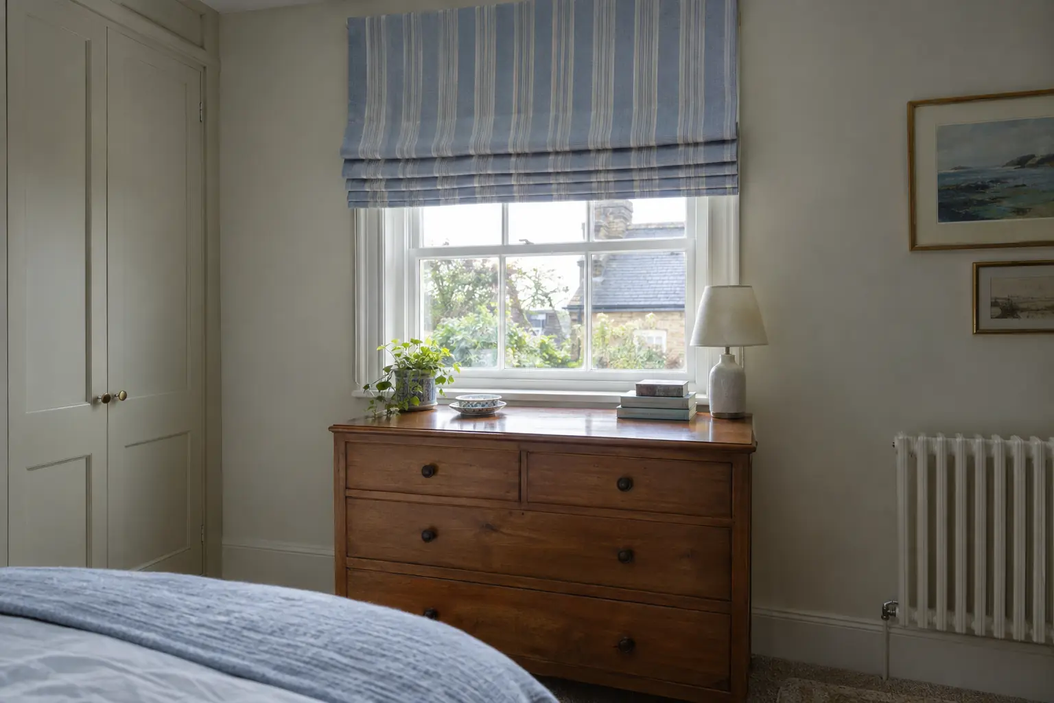 a roman blind in a soft blue stripe with a blackout lining raised into even folds above a small bedroom window, a chest of drawers pushed against the wall beneath the sill