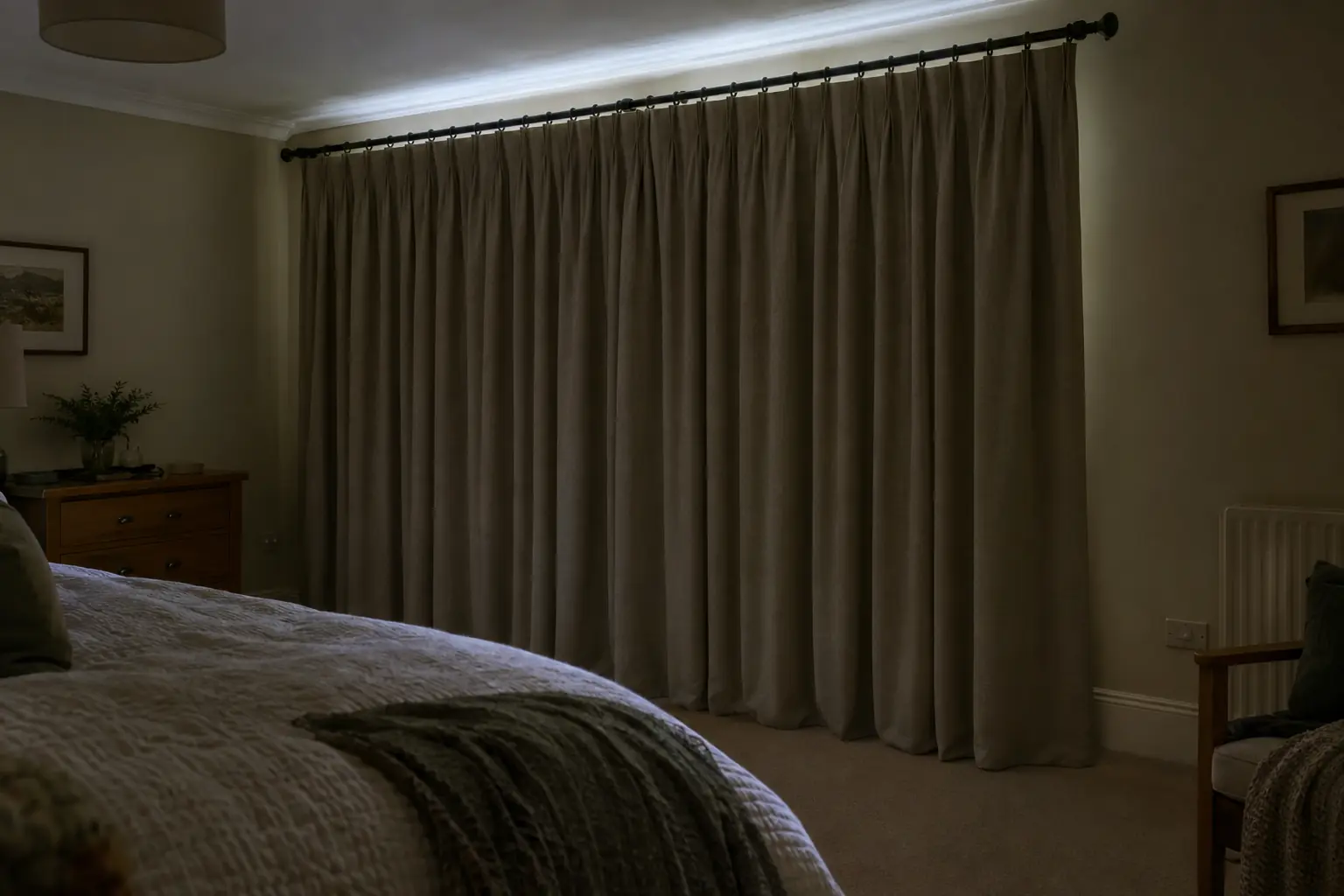 a bedroom in the early morning of midsummer, thick blackout-lined curtains drawn on a long pole across a wide window and hanging to the carpet, the room dim, a faint glow of daylight showing only above the pole on the ceiling