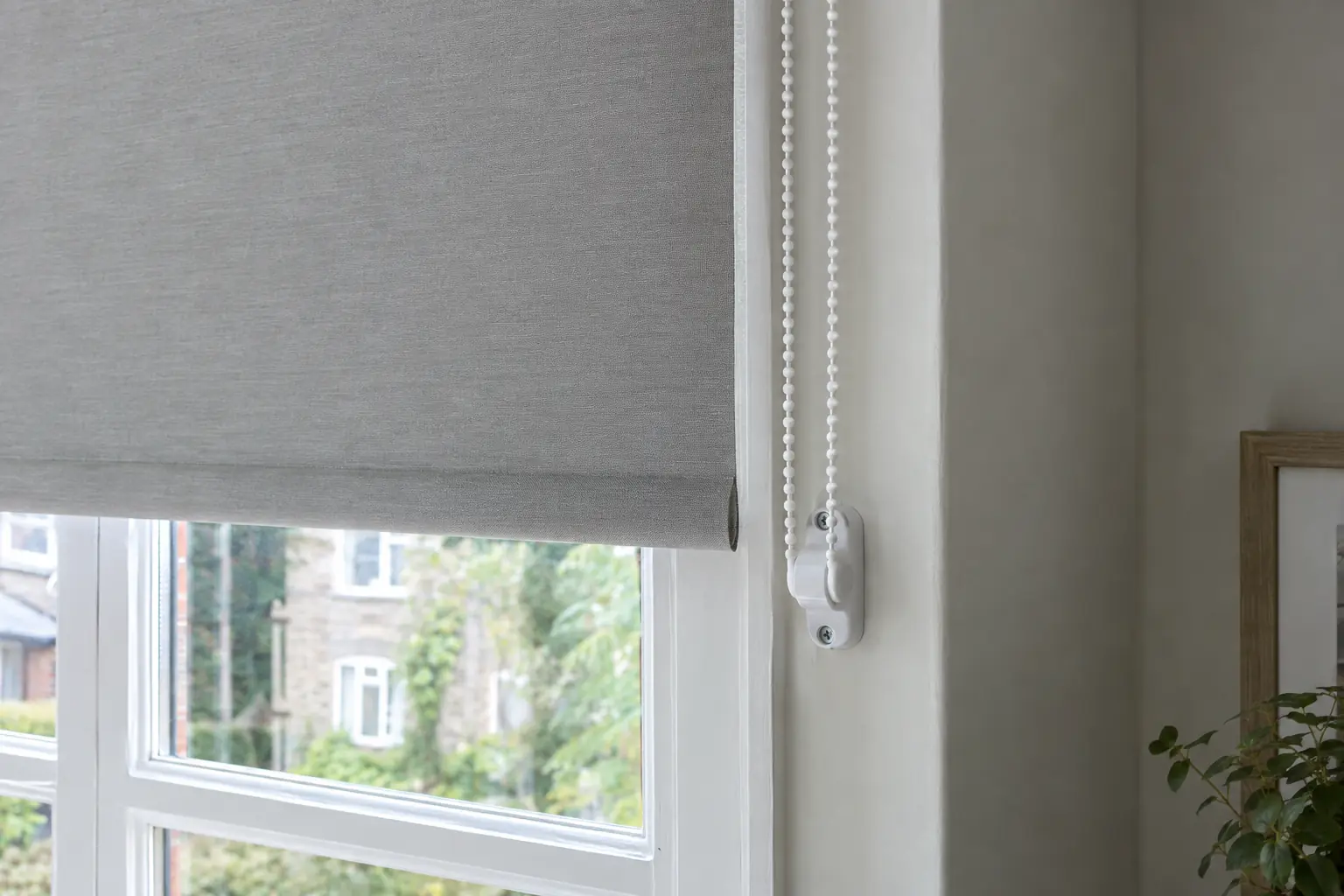 close view of a white plastic chain tensioner screwed to the side of a painted window reveal at shoulder height, holding the looped beaded chain of a roller blind taut against the wall