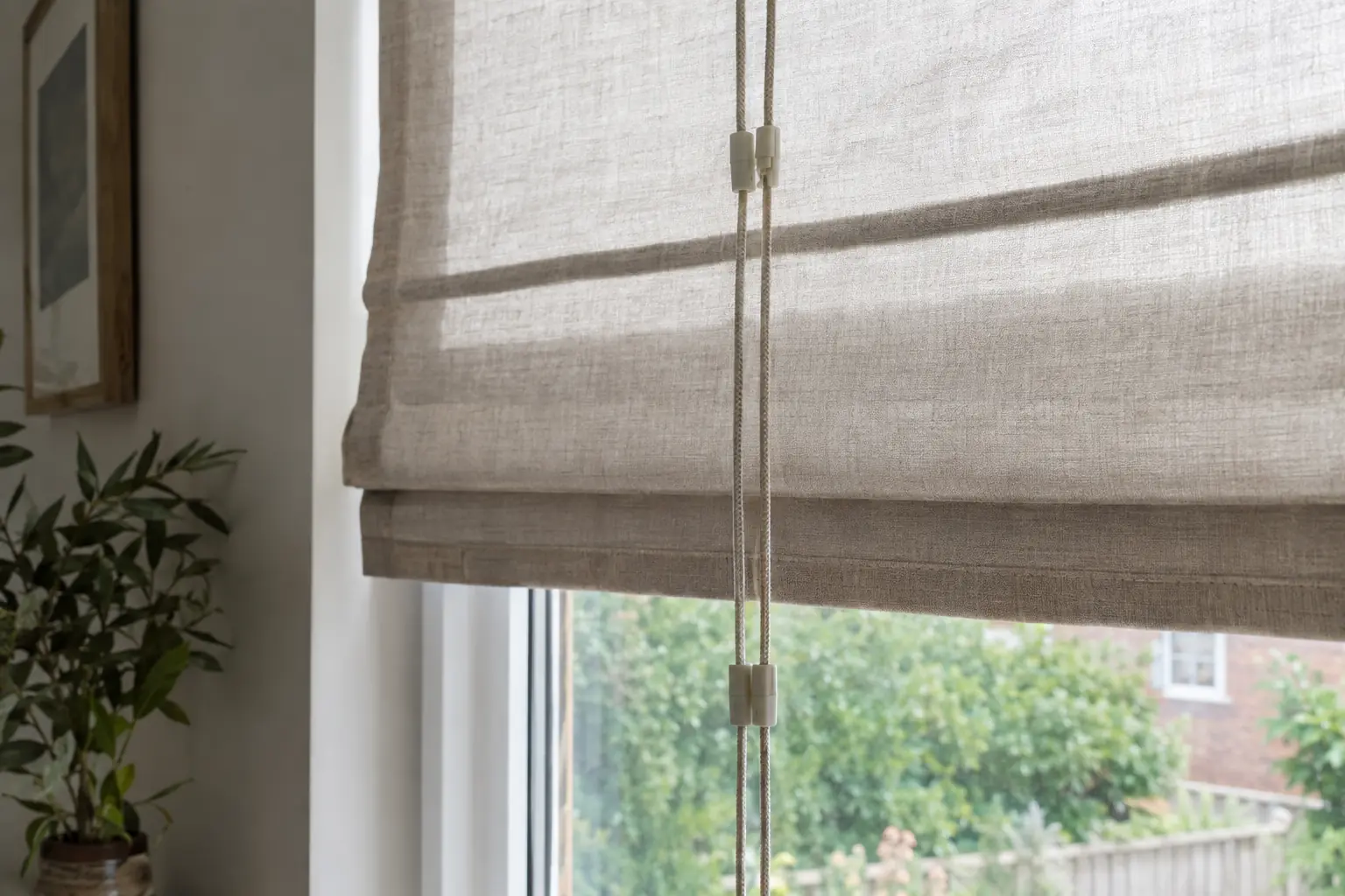 the back of a linen Roman blind seen from the window side, its vertical lifting cords joined by small plastic breakaway connectors, daylight coming through the glass behind