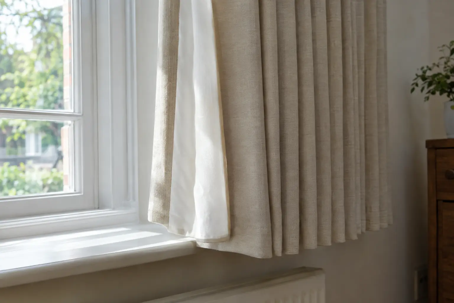 close view of the leading edge and hem of a cream lined pinch pleat curtain hanging just clear of a painted window sill, the pale lining showing where the sun falls on it