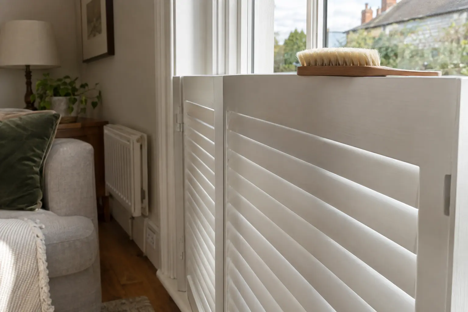 a close view of a soft-bristled brush resting on the top rail of a white painted hardwood shutter in a living room, the louvres closed flat and catching the light
