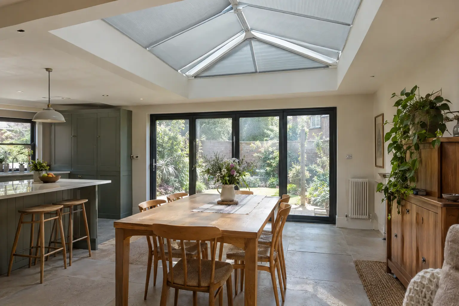 a flat-roofed kitchen extension with a rectangular roof lantern above the dining table, its pitched glass panels fitted with light grey tensioned blinds partly closed against midday sun