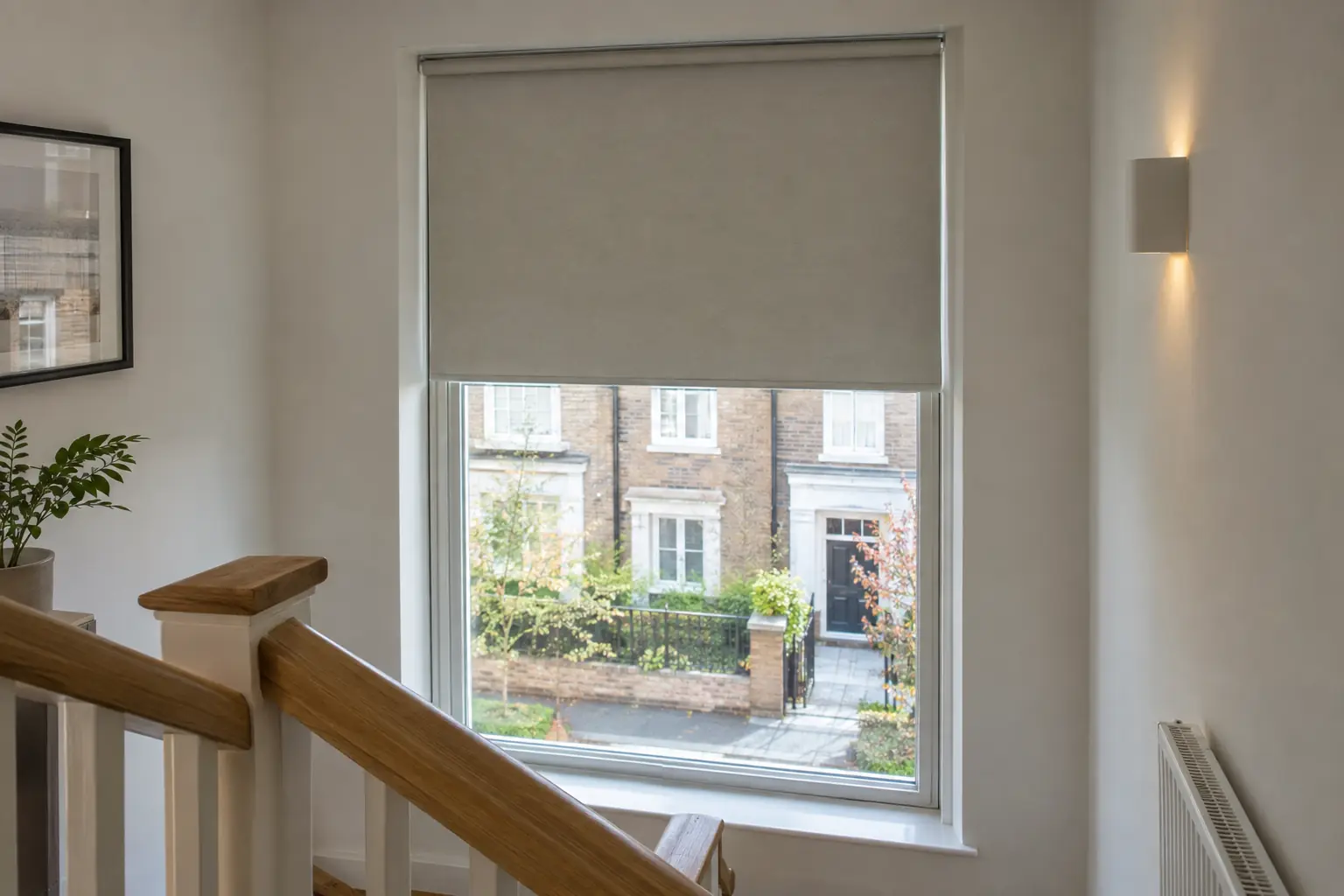 a tall stairwell window in a modern townhouse with a motorised roller blind half lowered over the glass, an oak stair handrail in the foreground and no cord or chain hanging at the side of the window