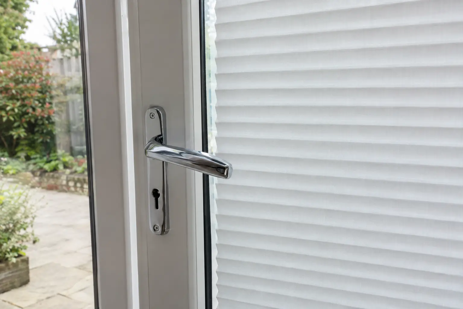 a close view of a chrome lever handle on an inward-opening French door, standing just in front of a white pleated blind fitted within the glazed panel, a narrow gap between the handle and the fabric