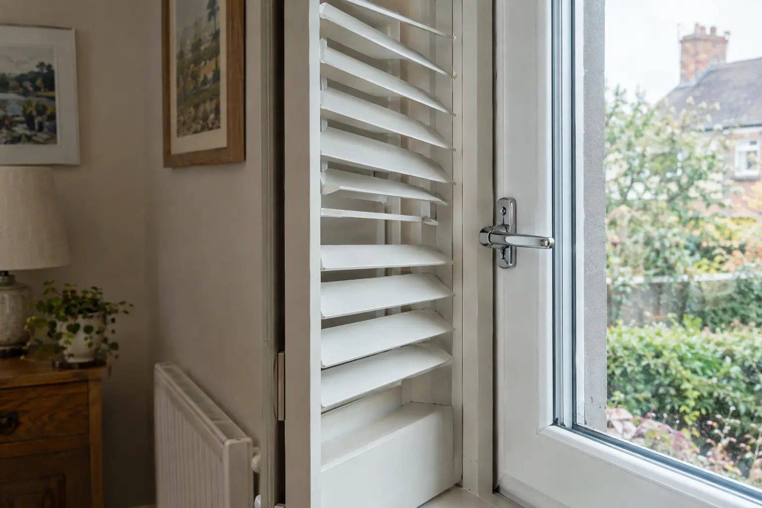 a close view of a shutter panel folded back against the side of a window reveal, the tilted louvres clearing a chrome lever handle on the window frame behind by a small margin