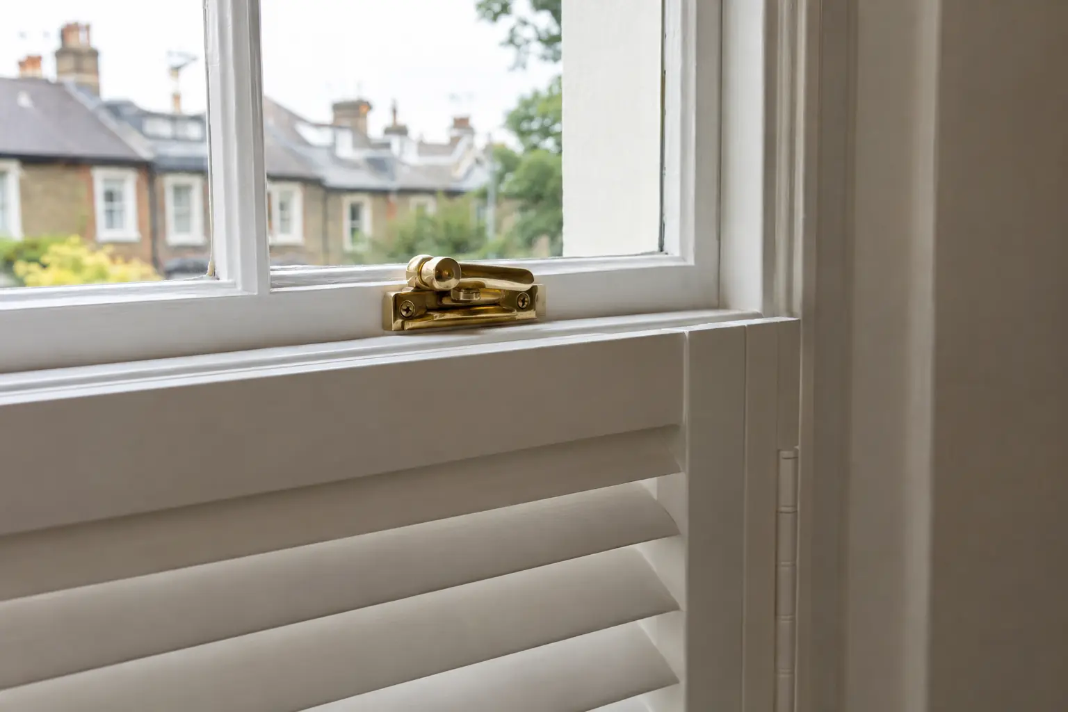 a close view of the meeting rail of a painted sash window with its brass catch, the top edge of a white café-style shutter lined up just below the rail