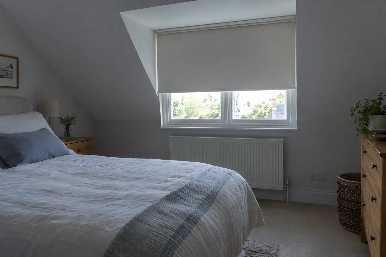 a loft bedroom under a sloping ceiling in summer, a pale blackout roller blind lowered on a dormer window, a light cotton bedspread on the bed and the room in cool shade while the sun is bright outside