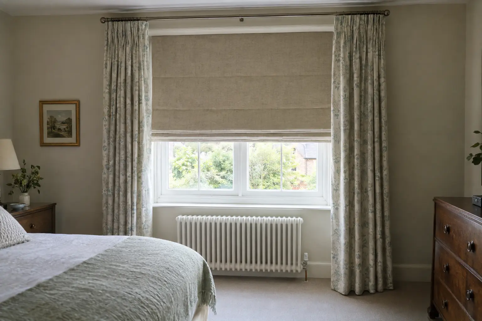 a bedroom window with a white radiator beneath it, a lined Roman blind lowered in the recess above the radiator and curtains left open at each side