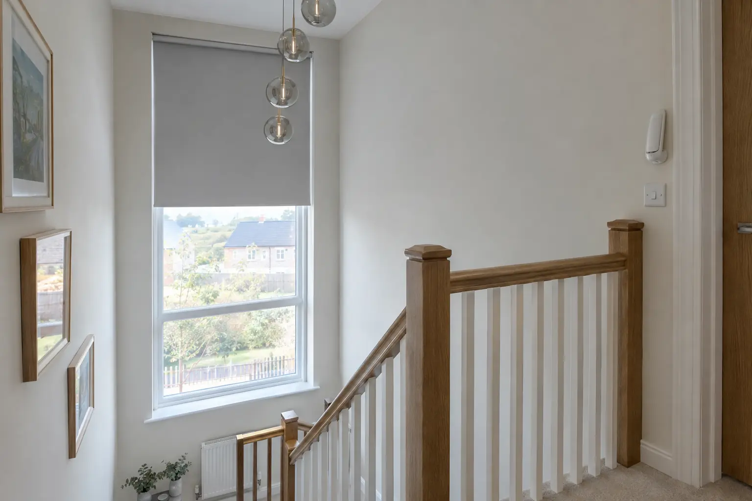 a double-height stairwell in a modern UK new-build with a tall landing window fitted with a pale grey motorised roller blind half lowered, a small white handset resting in a wall holder beside the light switch