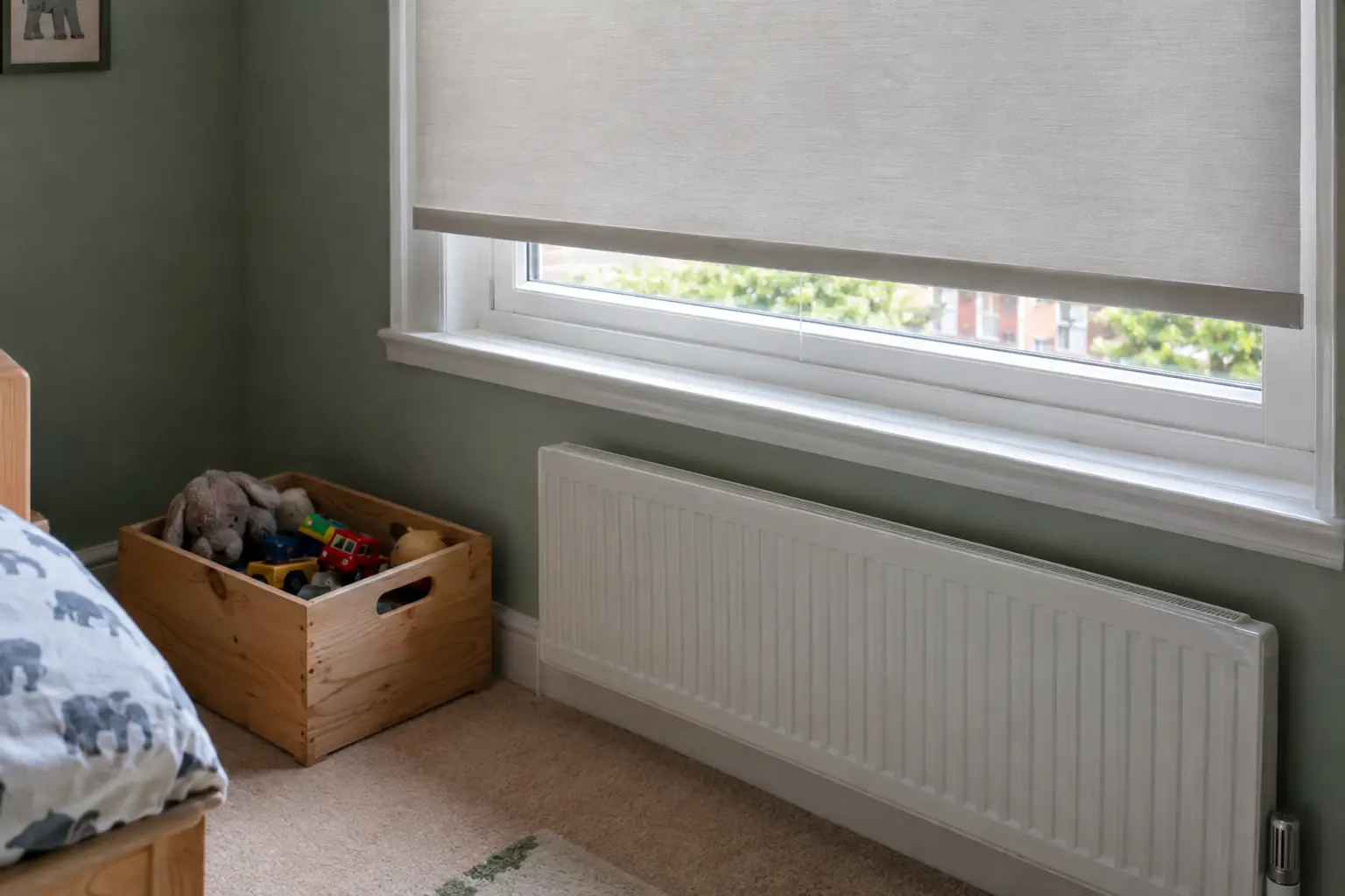 a close view of the bottom rail of a cordless blind in a child's bedroom, nothing hanging at either side of the window, a wooden toy box on the floor a little way from the wall