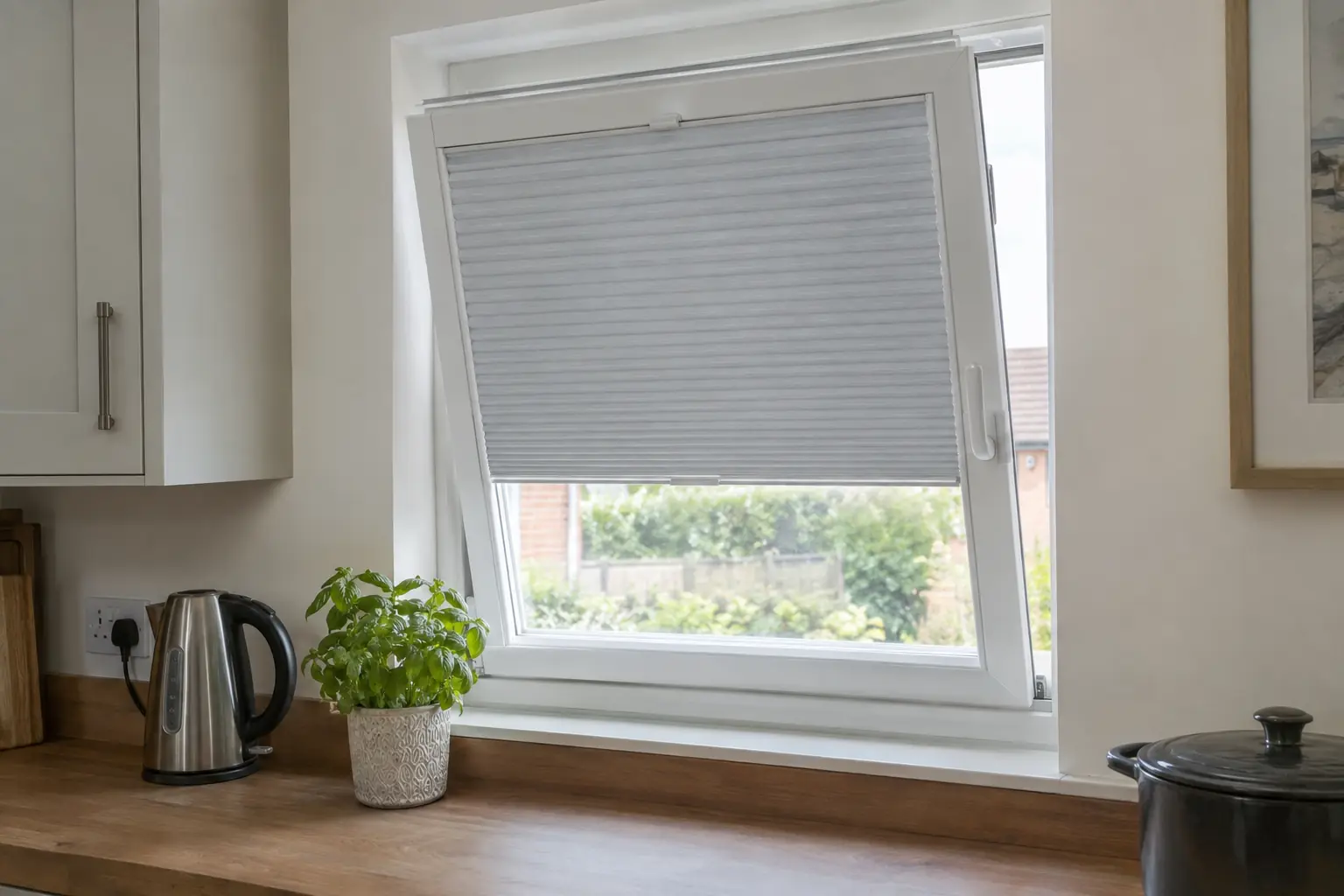 a white uPVC tilt and turn window in a modern kitchen, tipped open at the top for air, with a pale grey pleated blind in a slim white frame lying flat on the glass and tilting with the sash, a kettle and a pot of basil on the worktop beneath