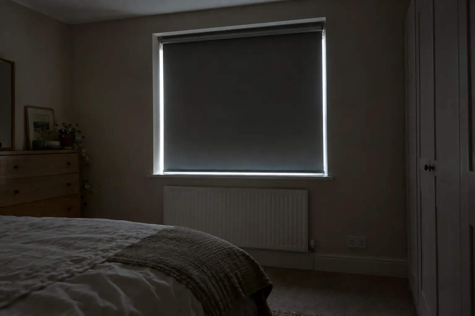 a dim bedroom at dawn with a blackout roller blind fitted inside the recess, a thin line of daylight showing down each side of the blind