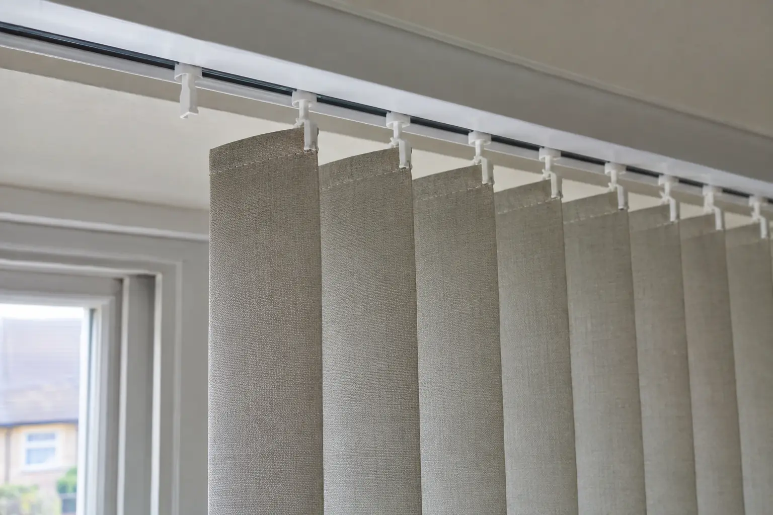 close view up into a vertical blind headrail showing a row of white plastic hanger clips gripping the tops of fabric louvres, one clip empty