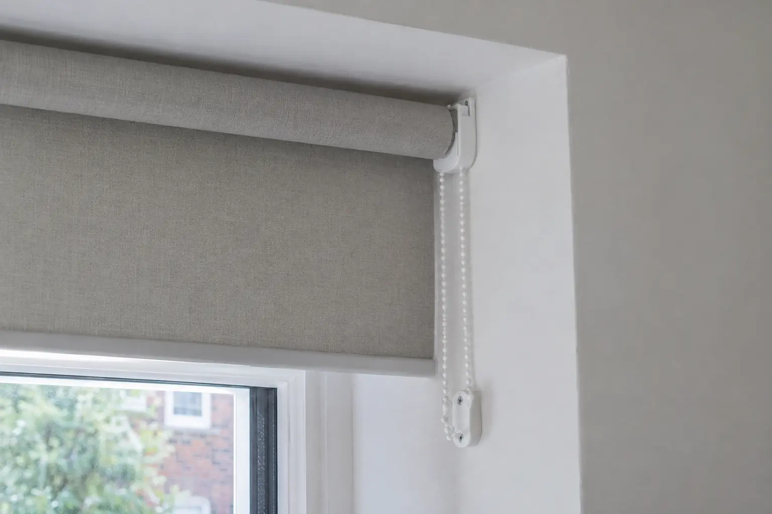 a close view of the top corner of a window recess showing the end of a roller blind: the fabric wound neatly on its tube, the white bracket, and a looped side chain held taut by a small child-safety device fixed to the plaster