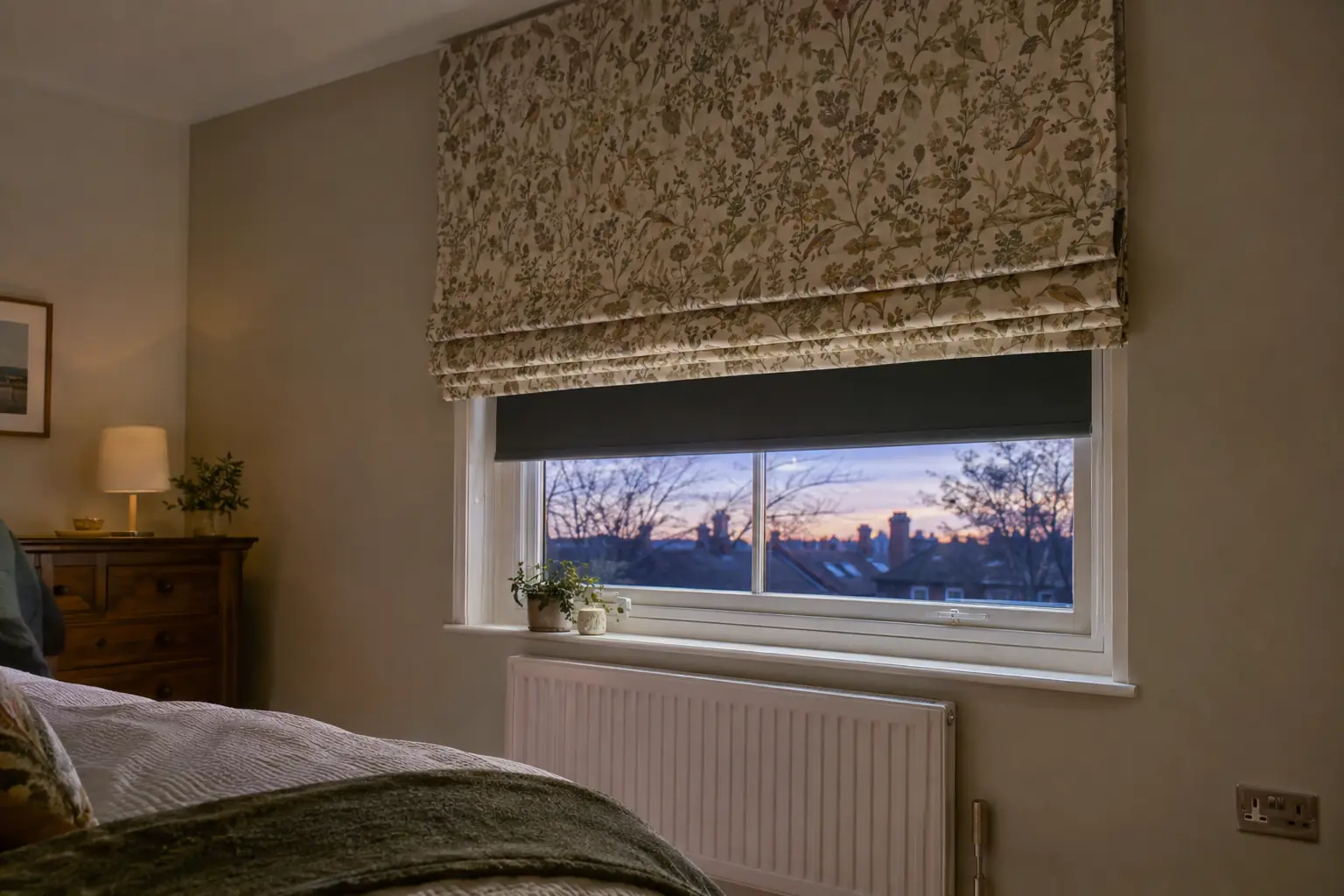 a bedroom window at dusk with a blackout roller blind fitted close to the glass inside the reveal and a patterned roman blind lowered in front of it on the wall