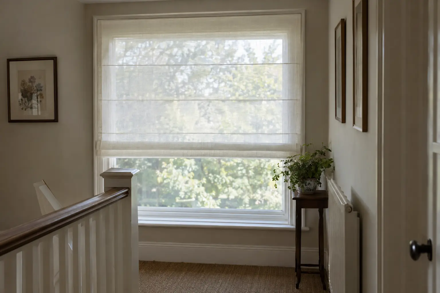 a sheer ivory voile roman blind lowered over a landing window in a 1930s house, the trees in the garden softened to a blur behind it in bright morning light