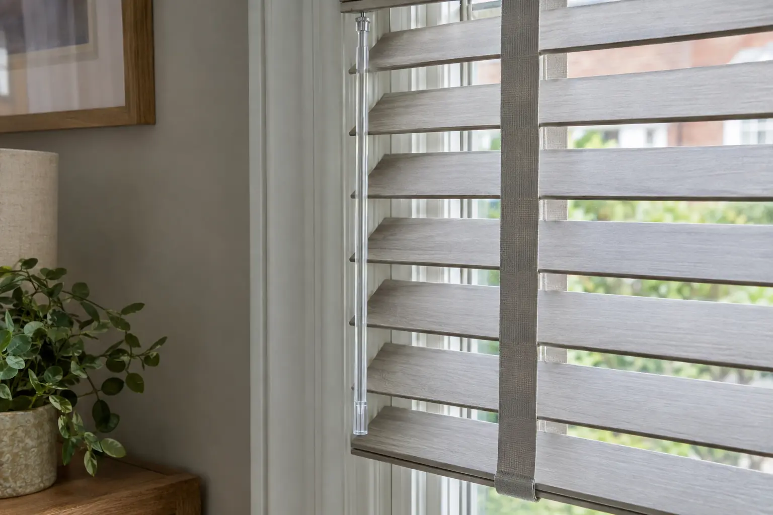 a close view of the edge of a soft grey faux wood venetian blind with cloth tapes running down over the ladder cords, a clear tilt wand hanging beside it against a painted window frame
