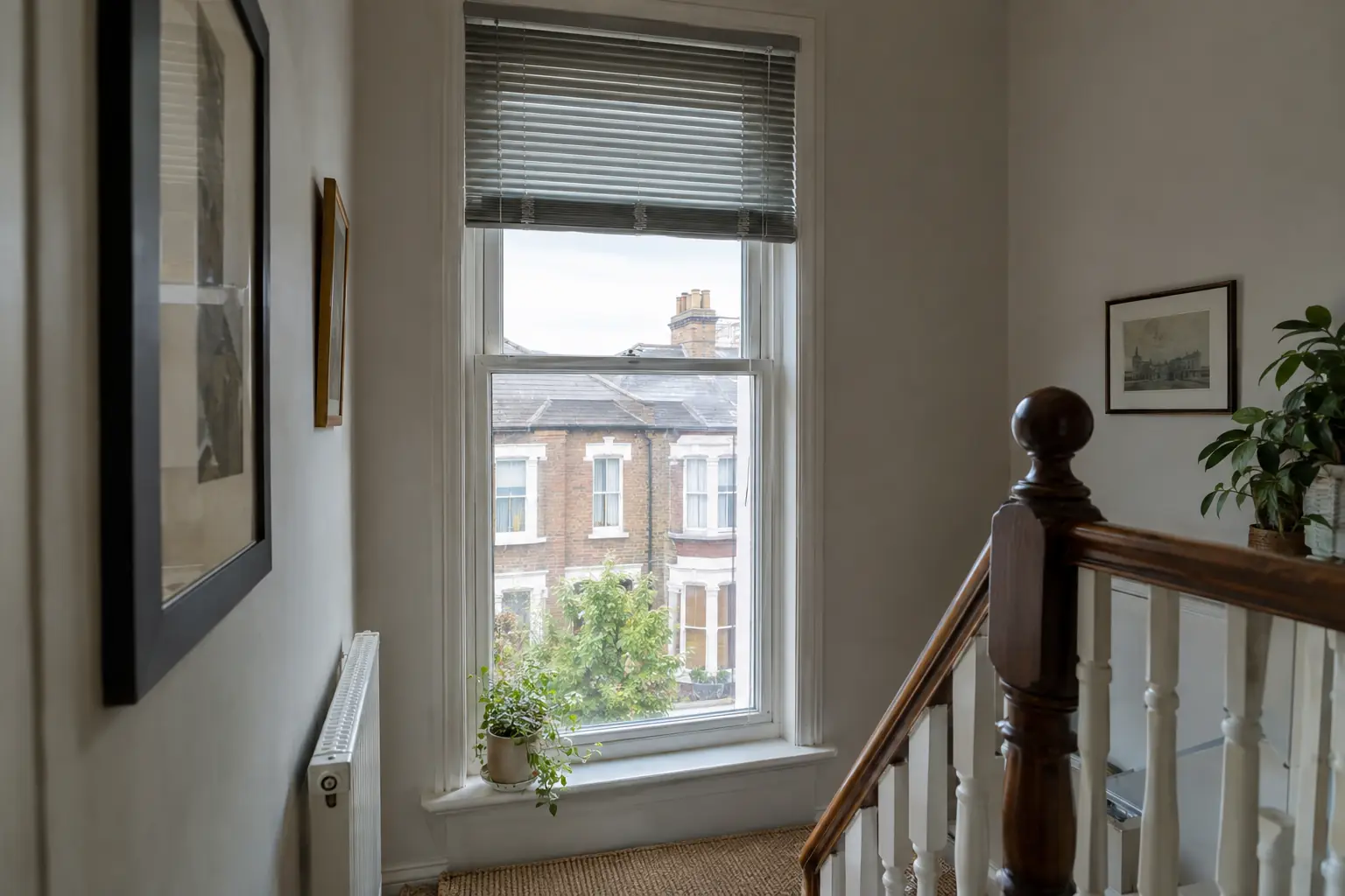 a tall narrow stairwell window in a Victorian terrace with a silver aluminium venetian raised halfway, its slats stacked neatly at the top and the rest of the glass clear