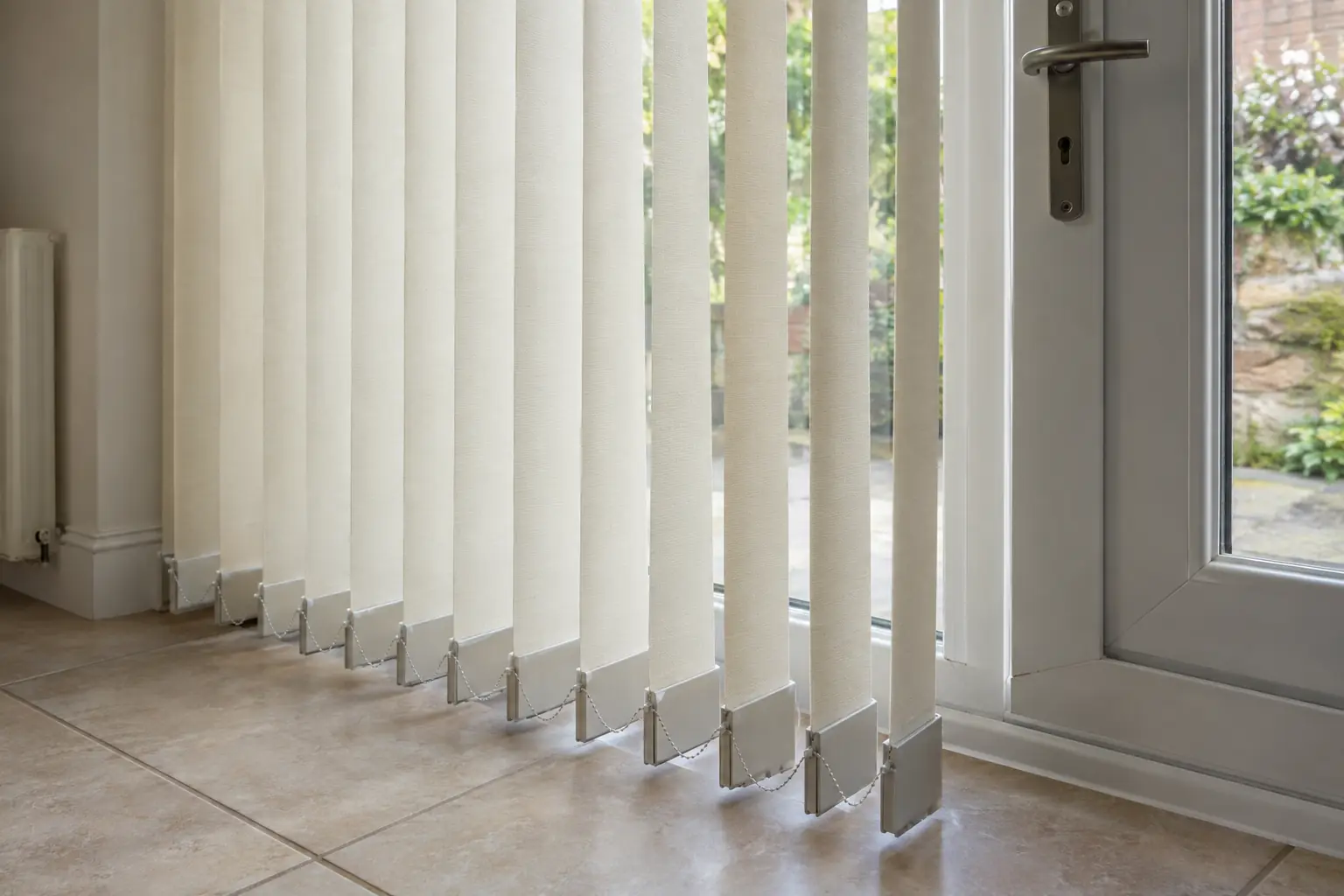 a close view of the bottom of cream fabric vertical louvres hanging over a tiled kitchen floor, each with its weight and a fine linking chain, the louvres turned partly open beside a door handle
