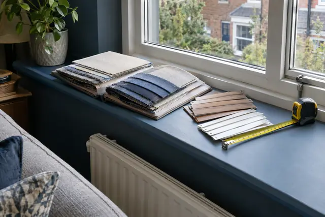 An open fabric sample book, wooden and white slat samples and a tape measure on a painted windowsill